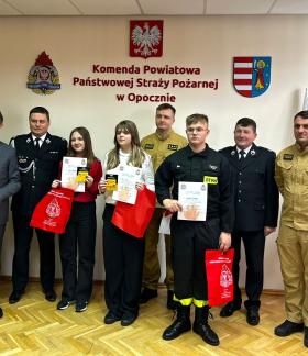 Osiem osób, w tym Starosta Opoczyński, strażacy i troje uczniów z dyplomami pozuje do zdjęcia w siedzibie PSP w Opocznie.  W tle ściana z nazwą jednostki PSP, godłem RP i herbami.