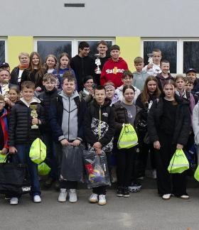 Duża grupa dzieci, młodzieży oraz dorosłych ustawiona w kilku rzędach przed budynkiem o jasnej elewacji z zielonymi elementami. Uczestnicy stoją blisko siebie i pozują do wspólnego zdjęcia. Wśród nich widoczni są uczniowie oraz opiekunowie, a także kilku umundurowanych policjantów