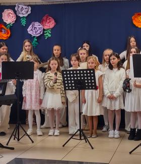 Grupa dzieci i młodzieży stoi na niewielkiej scenie podczas występu muzycznego w siedzibie PDPS w Drzewicy. Uczestnicy ubrani są głównie na biało. Na pierwszym planie kilka dziewczynek śpiewa do mikrofonu ustawionego na statywie. Obok znajdują się pulpity na nuty. Po lewej stronie osoba siedzi przy keyboardzie. W tle część uczestników gra na gitarach. Za nimi widoczna jest granatowa kurtyna ozdobiona dużymi, kolorowymi, papierowymi kwiatami.