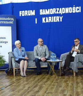 Na zdjęciu widoczny jest panel dyskusyjny podczas Forum Samorządności i Kariery w Szkole Podstawowej im. Jana Szymańskiego w Brzustowcu. Na sofie, fotelach bądź krzesłach siedzi sześć osób – czworo panelistów oraz dwie moderatorki po lewej stronie. Burmistrz Drzewicy trzyma mikrofon i zabiera głos. W tle znajduje się niebieska ścianka z napisem „Forum Samorządności i Kariery” oraz grafika przedstawiająca różne zawody. Po lewej stronie widoczny jest baner szkoły z portretem patrona.