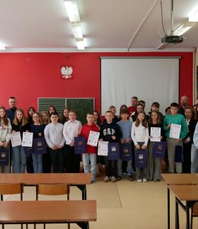 Na zdjęciu grupa uczniów, nauczycieli oraz członków komisji w sali lekcyjnej podczas podsumowania turnieju wiedzy o bezpieczeństwie w ruchu drogowym. Kilka osób trzyma w rękach dyplomy oraz granatowe torby z nadrukiem.