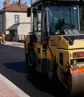 Modernizacja drogi z układaniem nowej nawierzchni asfaltowej przy użyciu specjalistycznych maszyn. Na zdjęciu widoczny walec drogowy oraz pracownicy w odzieży ostrzegawczej podczas prac budowlanych.