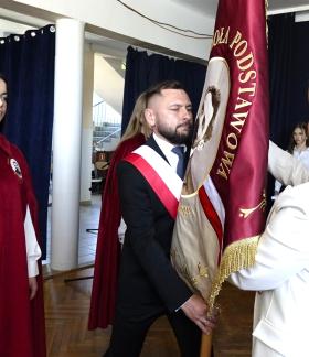 Wnętrze szkoły podczas uroczystości. Mężczyzna z biało-czerwoną szarfą trzyma sztandar szkoły w kolorze czerwonym z haftem. Naprzeciw stoi kobieta w jasnym stroju, poprawiająca lub przekazująca sztandar. Obok  uczennica w czerwonej pelerynie, patrząca w stronę sztandaru. 