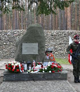 Uroczystość patriotyczna w lesie. Dwie żołnierki w mundurach polowych i bordowych beretach trzymają wartę z bronią po obu stronach pomnika-głazu z pamiątkową tablicą. U podnóża monumentu leżą liczne wieńce i znicze, a obok widoczne są polskie flagi. W tle znajduje się kamienny mur i wysoki las sosnowy.