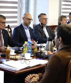 Przy stole z mikrofonami i dokumentami siedzi kilka osób w formalnych strojach – Burmistrz i Zastępca Burmistrza Drzewicy oraz radni. Głos zabiera Burmistrz Drzewicy, pozostali słuchają. Na stole widoczne są napoje i materiały do pracy.