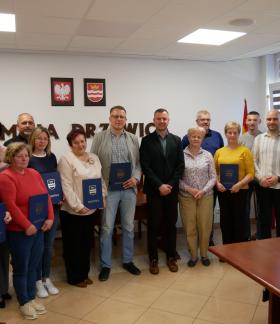 Zdjęcie grupowe przedstawicieli kół gospodyń oraz władz samorządowych w sali konferencyjnej Urzędu Miejskiego w Drzewicy. Uczestnicy trzymają pamiątkowe dyplomy w niebieskich okładkach z herbem gminy.