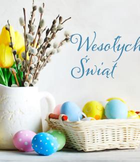 Na zdjęciu napis Wesołych Świąt, dzbanek z żółtymi tulipanami i baziami  obok koszyczek z jajkami oraz luźno leżące różnokolorowe  jajka