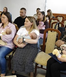 Grupa rodziców z niemowlętami podczas oficjalnego spotkania w Sali USC. W centralnej części siedzą trzy kobiety trzymające na rękach małe dzieci. W tle widoczni są pozostali uczestnicy siedzący na drewnianych krzesłach z jasną tapicerką.