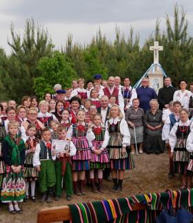 Duża grupa uczestników Majówki na ludowo w Gminie Drzewica, ubrana w barwne, tradycyjne stroje opoczyńskie, pozuje do pamiątkowego zdjęcia przy leśnej kapliczce w towarzystwie dzieci i kapłanów
