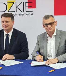 Członek Zarządu Województwa Łódzkiego Artur Bagieński i Burmistrz Drzewicy Janusz Reszelewski w garniturach siedzą przy stole z niebieskim obrusem i podpisują umowy. W tle widoczne logo „ŁÓDZKIE MARSZAŁKOWSKI” oraz fragment biało-czerwonej flagi.