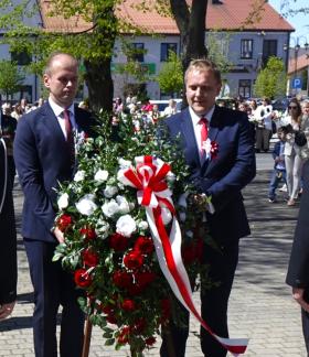 Dwóch urzędników w garniturach trzyma wieniec z biało-czerwonych kwiatów podczas uroczystości 3-majowych w parku na placu Wolności w Drzewicy. Obok stoją umundurowani strażacy, a w tle widoczny jest tłum uczestników oraz zabudowa miejska.