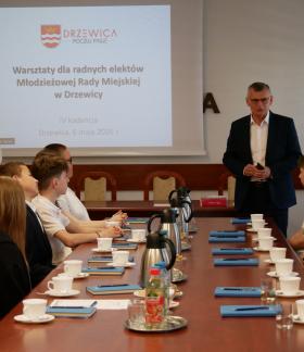 Na zdjęciu spotkanie warsztatowe: grupa młodzieży siedzi przy stole konferencyjnym, po prawej stronie stołu Burmistrza Drzewicy zabierający głos, po lewej Zastępca Burmistrza. Na ekranie wyświetlony jest slajd dotyczący warsztatów dla Młodzieżowej Rady Miejskiej w Drzewicy.