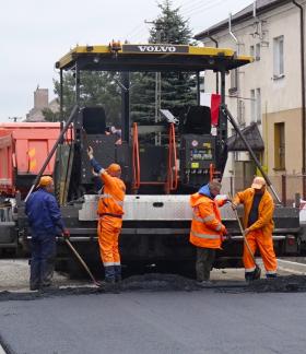 Prace drogowe na ulicy Braci Kobylańskich w terenie zabudowanym. Na środku duża maszyna do układania nawierzchni asfaltowej, za którą kilku pracowników w pomarańczowych strojach ochronnych rozprowadza świeżo ułożony asfalt. W tle samochody ciężarowe, budynki mieszkalne oraz drzewa. Pogoda pochmurna.