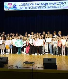 Na scenie sali widowiskowej RCK liczna grupa dzieci, młodzieży, opiekunów i samorządowców stoi w kilku rzędach przed ciemną kurtyną. Niektóre dzieci trzymają symboliczne czeki z nagrodami finansowymi oraz rekwizyty z wcześniejszych występów. Na górze duży biały transparent: „DRZEWICKI PRZEGLĄD TWÓRCZOŚCI ARTYSTYCZNEJ DZIECI I MŁODZIEŻY”. 