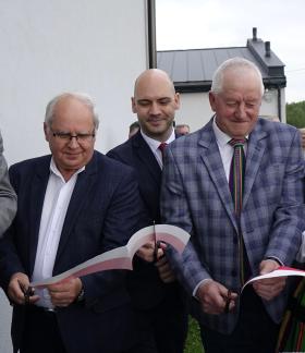  Zbliżenie na moment przecinania biało-czerwonej wstęgi na zewnątrz budynku. Wstęgę przecina pięć osób: Burmistrzowie Drzewicy, Prezes PKP Intercity Remtrak, Sołtys Idzikowic oraz Przewodnicząca KGW Idzikowice w tradycyjnym stroju ludowym.