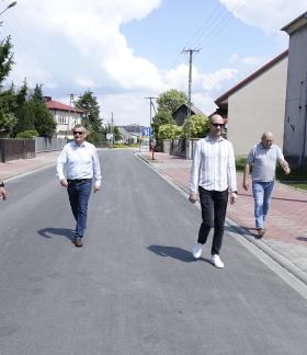 Pięciu mężczyzn idzie wzdłuż nowo wyremontowanej, asfaltowej ulicy w słoneczny dzień. Część idzie środkiem jezdni, pozostali po przylegającym do niej chodniku z kostki brukowej. W tle domy jednorodzinne, ogrodzenia oraz drzewa