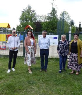 Oficjalne zdjęcie grupowe ośmiu osób stojących na trawie przed ogrodzonym, nowym placem zabaw z tablicą informacyjną w tle.