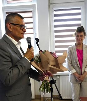 Burmistrz Drzewicy Janusz Reszelewski w szarym garniturze mówi do mikrofonu i przygotowuje się do wręczenia bukietu kwiatów stojącej naprzeciwko Skarbnik Gminy Irenie Jóźwickiej, ubranej w jasny garnitur i amarantową bluzkę.