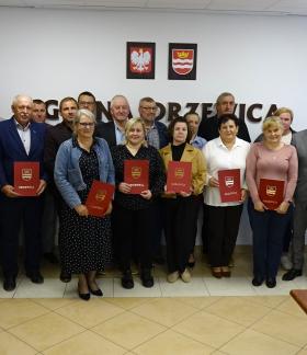  Ponad dwadzieścia osób pozuje do wspólnego zdjęcia w sali konferencyjnej Urzędu Miejskiego w Drzewicy. Część z nich trzyma w rękach czerwone teczki z herbem Drzewicy. W tle na białej ścianie duży napis „GMINA DRZEWICA” oraz godło Polski i herb gminy. Z prawej strony flagi: Polski i Unii Europejskiej.