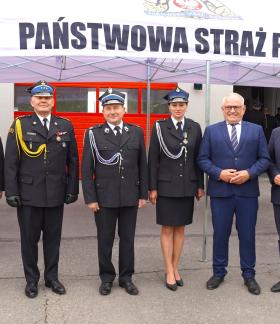Grupa sześciu osób stojących przed budynkiem pod białym namiotem z napisem „Państwowa Straż Pożarna”. Cztery osoby ubrane są w galowe mundury strażackie, dwie pozostałe w garnitury. W tle znajdują się czerwone bramy garażowe jednostki straży pożarnej. Uczestnicy stoją w jednym rzędzie i pozują do wspólnego zdjęcia.