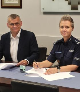  Podczas podpisywania porozumienia przy stole siedzą Burmistrz Drzewicy (w garniturze) i Pani Komendant Powiatowy Policji w Opocznie (w granatowym mundurze). Oboje trzymają długopisy i patrzą w stronę aparatu. Na stole znajdują się dokumenty oraz pieczątka. 