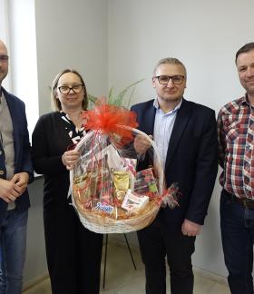 Cztery osoby – trzech mężczyzn i jedna kobieta – stoją obok siebie w jasnym pomieszczeniu biurowym. Pierwszy z lewej Zastępca Burmistrza Drzewicy, drugi od prawej Prezes PGKiM. Dwie osoby w centrum wspólnie trzymają duży, wiklinowy kosz prezentowy, wypełniony słodyczami i kawą