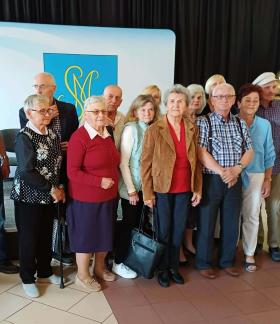 Grupowe zdjęcie kilkunastu seniorów, stojących blisko siebie w pomieszczeniu i pozujących do fotografii. Grupa składa się z kobiet i mężczyzn ubranych w stroje codzienne i wyjściowe. W tle czarna kotara, złote balony oraz jasna tablica z niebiesko-żółtym herbem przedstawiającym splecione litery. Po prawej stronie baner typu roll-up.