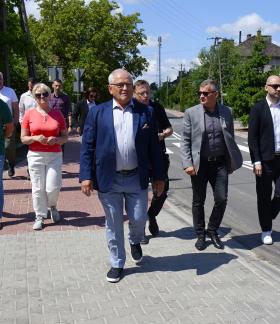 Oficjalna delegacja idzie w stronę obiektywu nowym chodnikiem z szarej kostki. W centrum I Wicewojewoda Łódzki w niebieskiej marynarce, otoczony przez innych uczestników spotkania.