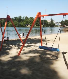 Nowoczesne huśtawki – tradycyjna oraz typu bocianie gniazdo – na pomarańczowych stelażach, ustawione na piaszczystej plaży nad jeziorem. W tle widoczni są kąpiący się i odpoczywający ludzie oraz zielony, przeciwległy brzeg.