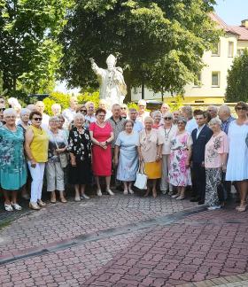 Liczna grupa uczestników spotkania pozuje do wspólnego zdjęcia na wybrukowanym placu kościelnym. W tle, za zebranymi ludźmi, widoczny jest biały pomnik przedstawiający papieża Jana Pawła II, otaczające go drzewa oraz żółty budynek mieszkalny.