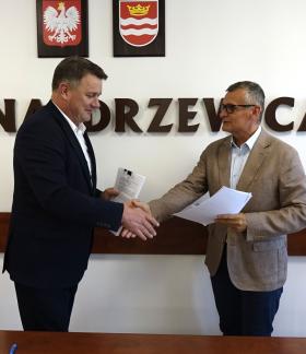 Członek Zarządu Woj. Łódzkiego Artur Bagieński i Burmistrz Drzewicy Janusz Reszelewski stoją w sali konferencyjnej i podają sobie dłonie w uścisku nad stołem, trzymając w rękach dokumenty. W tle biała ściana z dużym napisem „GMINA DRZEWICA”, nad którym wiszą godło Polski oraz herb Drzewicy.