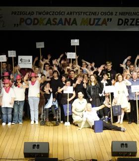 Na scenie w sali widowiskowej RCK kilkadziesiąt osób stoi obok siebie, uśmiechając się i machając rękami. W tłumie widoczne są osoby na wózkach inwalidzkich, osoby w różowych kapeluszach kowbojskich oraz trzymające tabliczki z nazwami różnych placówek (m.in. WTZ). Wysoko duży transparent festiwalu.