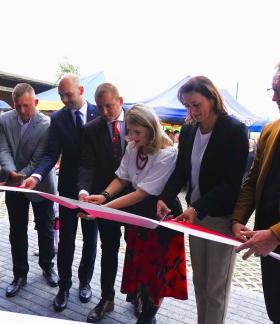 Oficjalny moment przecinania wstęgi. Grupa siedmiu osób – sześciu mężczyzn w garniturach lub marynarkach oraz dwie kobiety – stoi w rzędzie na zewnątrz budynku. Wspólnie trzymają i przecinają nożyczkami biało-czerwoną wstęgę. W tle widoczne są namioty festynowe oraz fragmenty otoczenia.