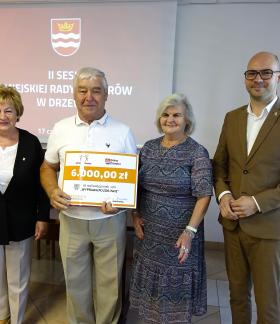 Pozowane zdjęcie czterech osób stojących obok siebie w sali konferencyjnej. Drugi od lewej mężczyzna trzyma duży, symboliczny czek na kwotę 6000,00 zł na realizację projektu „WYPRAWA PO ZDROWIE”. Po prawej stronie stoi mężczyzna w jasnobrązowym garniturze i okularach. W tle widoczny jest ekran projekcyjny z napisem „II SESJA MIEJSKIEJ RADY SENIORÓW W DRZEWICY” oraz flagi państwowe.