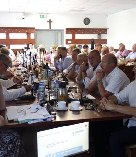Szeroka panorama sali konferencyjnej Urzędu Miejskiego w Drzewicy. Przy długim stole siedzi kilkunastu mężczyzn i kobiet ubranych oficjalnie. Uczestnicy sesji, w tym Burmistrz Janusz Reszelewski, skupieni są na dokumentach i ekranie, na którym widoczny jest porządek obrad XXV sesji Rady Miejskiej. Na stole znajdują się mikrofony, butelki z wodą i filiżanki.