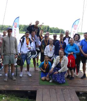 Grupa kilkunastu osób w różnym wieku (młodzież i dorośli) pozuje do wspólnego zdjęcia na drewnianym pomoście nad wodą. Część osób ma na szyi medale, a w tle widać flagi reklamowe z herbem Drzewicy. Wszyscy są w letnich, sportowych strojach. Na pierwszym planie po prawej stronie stoi Burmistrz Drzewicy w ciemnej marynarce i jasnej koszuli, a obok niego, na środku pomostu, klęczy kobieta w niebieskiej spódnicy w kwiaty oraz młoda dziewczynka w sportowym stroju.