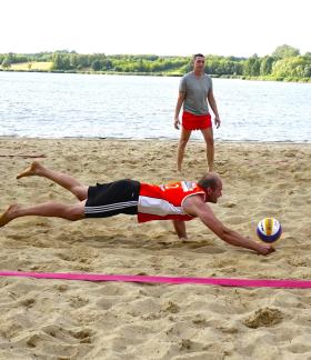 Siatkarz plażowy w pomarańczowej koszulce wykonuje widowiskowy rzut, rzucając się na piasek w locie, aby podbić piłkę tuż nad ziemią. W tle kolega z drużyny i jezioro.