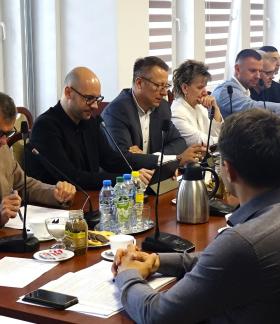 Uczestnicy sesji Rady Miejskiej w Drzewicy siedzą po obu stronach stołu konferencyjnego i biorą udział w obradach. Pierwszy z lewej Burmistrz Drzewicy odczytuje dokument. Na stołach znajdują się dokumenty, mikrofony oraz napoje.