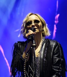 Anna Wyszkoni podczas koncertu śpiewa do mikrofonu na oświetlonej niebiesko-fioletowym światłem scenie. Artystka ma krótkie blond włosy, ciemne okulary i czarną skórzaną kurtkę z frędzlami. W tle widoczny jest ekran LED