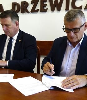 Członek Zarządu Woj. Łódzkiego Artur Bagieński i Burmistrz Drzewicy Janusz Reszelewski w garniturach siedzą obok siebie przy drewnianym konferencyjnym stole. Obaj podpisują leżące przed nimi dokumenty. Na ścianie w tle widoczny duży, brązowy, wypukły napis „GMINA DRZEWICA”.
