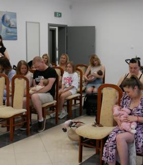 Burmistrz Drzewicy przemawia do rodzin zgromadzonych w jasnej sali urzędowej. Po lewej stronie, na stole, ułożone są przygotowane upominki w ozdobnych pudełkach z pluszowymi misiami. W rzędach krzeseł siedzą rodzice trzymający na rękach noworodki i małe dzieci.