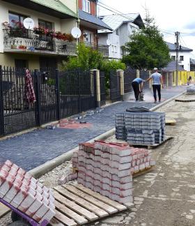 a osiedlowej ulicy trwają prace związane z przebudową drogi. Widoczne są nowo ułożone chodniki z kostki brukowej, krawężniki, materiały budowlane oraz pracownicy wykonujący roboty. W tle znajdują się domy jednorodzinne.