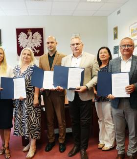 Grupa ośmiu osób w gabinecie pozuje do wspólnego zdjęcia. Cztery osoby prezentują otwarte niebieskie teczki z podpisanymi dokumentami. W środku Zastępca Burmistrza Drzewicy Dominik Niemirski.