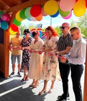 roczyste przecięcie czerwonej wstęgi pod drewnianą wiatą w Domasznie, przyozdobioną kolorowymi balonami podczas Pikniku Rodzinnego.