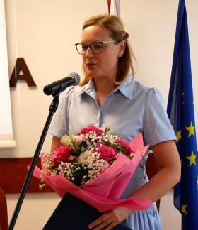 ani Dyrektor Przedszkola Samorządowego w Drzewicy w okularach i błękitnej sukience stoi przy mikrofonie, trzymając w rękach duży bukiet różowych i białych kwiatów oraz niebieską teczkę. Kadr prezentuje ujęcie od pasa w górę. W tle widać flagę Unii Europejskiej oraz ekran projekcyjny.