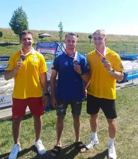 Trzej uśmiechnięci kajakarze górscy LKK Drzewica - od lewej: Szymon Zawadzki, Michał Franczak, Maciej Bartos - stoją na trawie przy torze kajakowym i prezentują zdobyte medale. Dwaj z nich mają na sobie żółte koszulki polo, a środkowy – niebieską koszulkę polo.