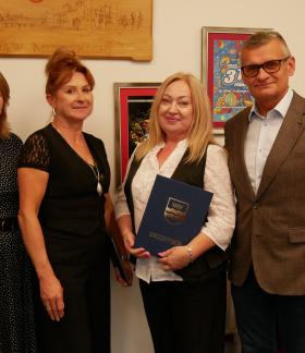 Pozowane zdjęcie w gabinecie Burmistrza Drzewicy. Od lewej trzy panie -  Dyrektor ZEAS, Dyrektor SP w Domasznie, Dyrektor SP w Domasznie – oraz Burmistrz Drzewicy. Na ścianie za nimi wiszą oprawione plakaty 31. i 32. Finału WOŚP oraz rzeźbiona drewniana tablica.