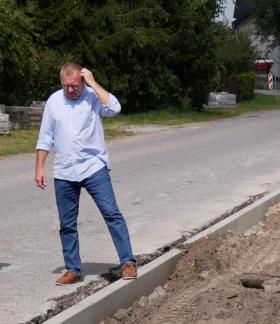 Trzech mężczyzn w strojach wizytowych ogląda plac budowy drogi ze świeżo ułożonymi krawężnikami na pierwszym planie oraz pracującą w oddali koparką