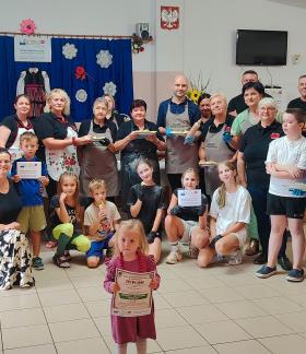  Uczestnicy i organizatorzy warsztatów lepienia pierogów pozują do pamiątkowego zdjęcia na świetlicy wiejskiej w Zakościelu. Część osób trzyma dyplomy i talerze z gotowymi pierogami, a na pierwszym planie mała dziewczynka dumna prezentuje oprawiony dyplom za udział w warsztatach.