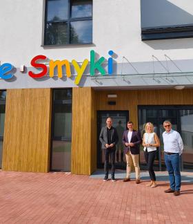 Cztery osoby stoją przed nowoczesnym budynkiem z kolorowym napisem „Wesołe Smyki” nad wejściem.